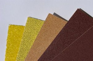 Fächerförmig angeordnete Schleifpapierbögen in verschiedenen Farben und Körnungen, von grob bis fein. Die Körnungen reichen von gelbem grobkörnigem Papier bis zu feinem dunkelbraunem Schleifpapier, dargestellt auf weißem Untergrund.