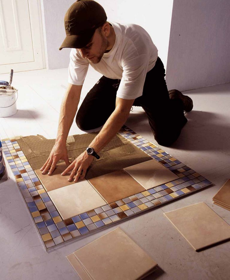 Fliesenleger verlegt Bodenfliesen auf frischem Fliesenkleberbett, umgeben von einem Mosaikrahmen mit blauen, braunen und weißen Fliesen.