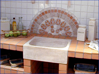 Alt-Text: Küchenspüle mit Arbeitsfläche aus rötlichem Naturstein-Mosaik, Wandverkleidung mit weiß glasierten Fliesen und dekorativem Mosaikbogen in Orange- und Beigetönen. Auf der Arbeitsfläche stehen Vorratsbehälter, eine Obstschale und Ölflaschen.