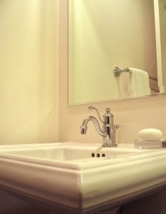 Ein Badezimmer mit einem weißen Waschbecken, einem verchromten Wasserhahn und einem darüber hängenden rechteckigen Spiegel. Rechts im Bild ist ein Handtuch auf einem Handtuchhalter zu sehen und daneben liegt eine Seife auf einer Ablage.