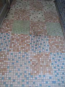Mosaikboden mit quadratischen Fliesen in verschiedenen Farben, darunter Blau, Grün, Beige und Weiß. Die Fliesen sind in einem geometrischen Muster angeordnet. Einige Bereiche zeigen Abnutzung oder Schmutz.