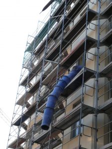 Mehrstöckiges Baugerüst mit Bauschuttabwurfschacht an einem Wohngebäude. Der blaue Schuttkanal ragt aus dem oberen Stockwerk und ist an der Fassade befestigt.