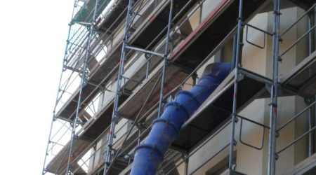 Mehrstöckiges Baugerüst mit Bauschuttabwurfschacht an einem Wohngebäude. Der blaue Schuttkanal ragt aus dem oberen Stockwerk und ist an der Fassade befestigt.