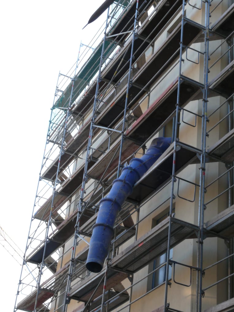 Mehrstöckiges Baugerüst mit Bauschuttabwurfschacht an einem Wohngebäude. Der blaue Schuttkanal ragt aus dem oberen Stockwerk und ist an der Fassade befestigt.