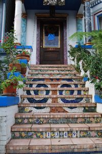 Bunte Freitreppe mit kunstvoll verzierten Mosaikfliesen in floralen und geometrischen Mustern, die zu einem Eingang mit Holztür und üppiger Pflanzen-Dekoration führt; Hausnummer 1349 am untersten Treppenabsatz.