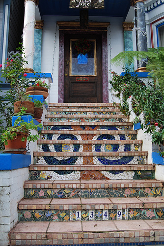 Bunte Freitreppe mit kunstvoll verzierten Mosaikfliesen in floralen und geometrischen Mustern, die zu einem Eingang mit Holztür und üppiger Pflanzen-Dekoration führt; Hausnummer 1349 am untersten Treppenabsatz.