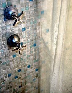 Zwei Metall-Wasserhähne an einer Wand mit Mosaikfliesen in Blau- und Grüntönen. Rechts ist ein durchsichtiger Duschvorhang zu sehen, der teilweise die Sicht verdeckt. Die Szene zeigt einen klassischen Duschbereich.