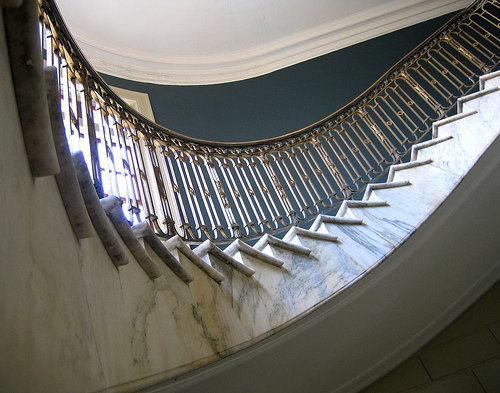 Elegante gewendelte Natursteintreppe aus poliertem Marmor mit kunstvoll verziertem Geländer aus Metall in einem klassischen Gebäude – Beispiel für hochwertige Natursteintreppen im Innenbereich.