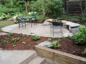 Mit Steinplatten gepflasterte Terrasse im Garten mit schwarzem Metalltisch und vier passenden Stühlen. Die Terrasse ist von neu angelegten Beeten mit frischer Rindenmulchschicht umgeben. Im Hintergrund stehen Sträucher, Hortensien und ein Holzzaun, links führen Treppenstufen zur Terrasse hinauf.