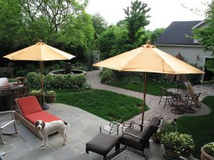 Eine Terrasse mit zwei gelben Sonnenschirmen, einem roten Liegestuhl und einem Tisch mit Stühlen. Ein Hund steht auf den Fliesen. Im Hintergrund ein Garten mit Gehweg, Rasen und Pflanzen, sowie ein Nebengebäude mit grauem Dach.