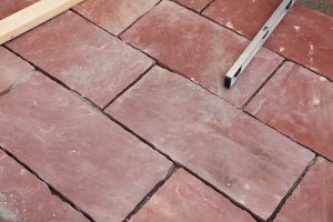 Verlegte rote Natursteinplatten mit unregelmäßigen Fugen auf einem Boden einer Terrasse. Ein Wasserwaage und ein Holzbrett liegen zur Ausrichtung der Platten daneben.