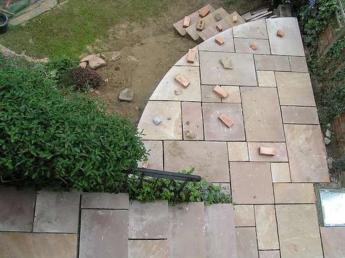 Blick von oben auf eine im Bau befindliche Naturstein-Terrasse mit hellen, rechteckigen Platten in verschiedenen Größen. Auf der Fläche liegen verstreut einige rote Ziegelsteine. Links führen Natursteinstufen mit schwarzem Handlauf hinab zur Terrasse, rechts ist eine mit Efeu bewachsene Mauer sichtbar.