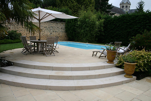 Eine Terrasse mit runden Stufen führt zu einem kleinen Pool. Auf der Terrasse stehen Holzmöbel und ein Sonnenschirm. Im Hintergrund sind eine Steinmauer, dichtes Grün und ein Kirchturm sichtbar.