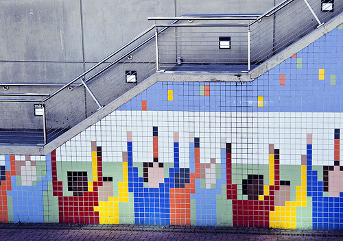 Wandbild aus bunten Fliesen an einer Treppe, das Menschen mit erhobenen Armen in Pixelkunst darstellt. Die Figuren tragen verschiedene farbige Kleidung. Die Wand ist mit einem Metallgeländer gesichert.