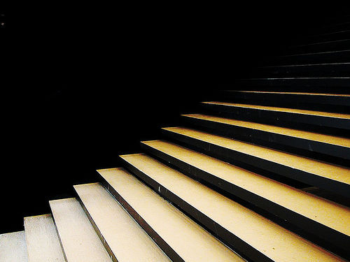 Schwarzweiß-Foto von minimalistischen Treppenstufen, die diagonal von links unten nach rechts oben verlaufen. Die Treppen sind hell beleuchtet und heben sich stark vom dunklen Hintergrund ab, was einen dramatischen Kontrast erzeugt.
