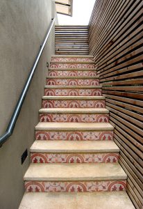 Freitreppe mit dekorativen, gemusterten Fliesen auf den Setzstufen und schlichten Trittflächen, flankiert von einer modernen Holzlattung und einem schlichten Metallhandlauf – stilvolles Beispiel für gemusterte Außentreppen im mediterranen Design.