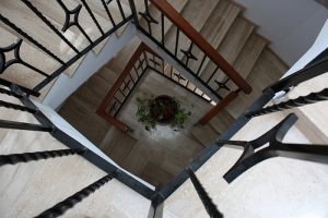 Blick von oben auf ein Treppenhaus mit hellen, polierten Natursteinstufen und schwarzem Metallgeländer mit Holzhandlauf – modernes Design mit robustem Natursteinbelag. In der Mitte des Treppenhauses steht eine dekorative Zimmerpflanze.