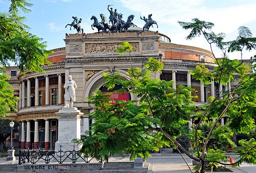 Gebäude mit reich verzierter Fassade und einer Skulpturengruppe mit Reitern auf dem Dach. Im Vordergrund eine Statue und grüne Bäume, die den Platz umgeben. Klarer Himmel und Tageslicht betonen die architektonischen Details der Szenerie.