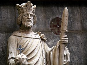 Sandsteinfigur eines Königs aus Ruhrsandstein vor einer Steinwand. Die Figur trägt eine Krone, hält ein Zepter in der rechten Hand und einen Reichsapfel in der linken Hand.