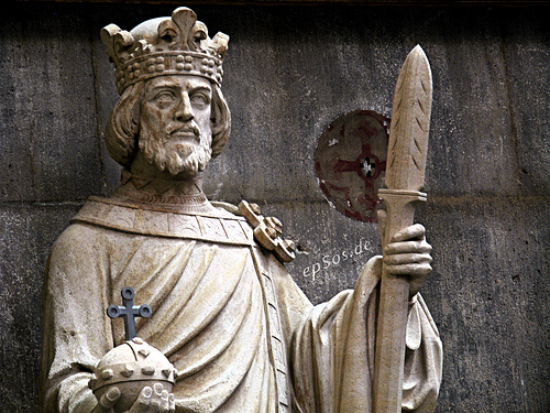 Sandsteinfigur eines Königs aus Ruhrsandstein vor einer Steinwand. Die Figur trägt eine Krone, hält ein Zepter in der rechten Hand und einen Reichsapfel in der linken Hand.