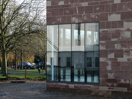 Sandsteinfassade eines modernen Gebäudes mit großflächigen Platten aus Naturstein in rötlich-beigen Tönen, die unregelmäßig angeordnet sind. In der Fassade befinden sich große Fenster mit Blick ins helle Treppenhaus, im Hintergrund sind Bäume und parkende Autos zu sehen.
