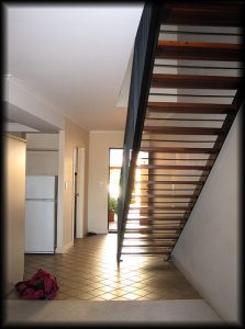 Ein moderner, offener Wohnbereich mit hellen Fliesen, einer Holztreppe mit Metallgeländer und einem weißen Kühlschrank links. Ein geöffneter Eingangsbereich mit Licht von draußen ist sichtbar. Eine rosa Tasche liegt auf dem Boden.