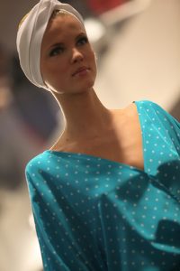 Eine Person auf einem Laufsteg trägt ein türkisfarbenes, gepunktetes Kleid mit tiefem Ausschnitt und ein weißes Kopfband. Der Hintergrund ist unscharf, was den Fokus auf die modische Kleidung lenkt. Die Beleuchtung ist weich.