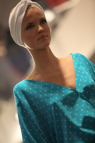 Eine Person auf einem Laufsteg trägt ein türkisfarbenes, gepunktetes Kleid mit tiefem Ausschnitt und ein weißes Kopfband. Der Hintergrund ist unscharf, was den Fokus auf die modische Kleidung lenkt. Die Beleuchtung ist weich.