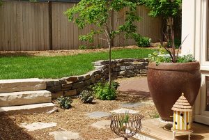 Gartenbereich mit kleiner Natursteinmauer aus indischem Sandstein, bepflanzt mit Sträuchern und einem jungen Baum, daneben ein großer Terrakottatopf und ein dekoratives Vogelhäuschen auf einer Holzstufe.