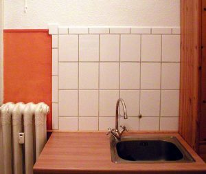 Küchenzeile mit weiß gefliester Rückwand, Spülbecken mit hoher Armatur, links ein Rippenheizkörper, rechts eine hölzerne Küchenschrankseite.