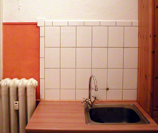 Küchenzeile mit weiß gefliester Rückwand, Spülbecken mit hoher Armatur, links ein Rippenheizkörper, rechts eine hölzerne Küchenschrankseite.
