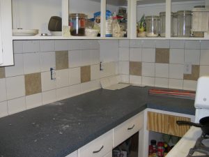 Küchenarbeitsbereich während der Renovierung: Teilweise geflieste Rückwand mit weißen und beigen Fliesen, einige Steckdosen noch ohne Abdeckung, Werkzeuge und Mörtelreste auf der dunklen Arbeitsplatte, offene Schränke mit Lebensmitteln und Küchenutensilien im Hintergrund.