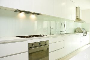 Moderne, minimalistische Einbauküche mit hochglänzendem, durchgehendem Fliesenspiegel aus Glas in Weiß, der sich über die gesamte Wandfläche hinter Arbeitsplatte, Spüle und Kochfeld erstreckt.