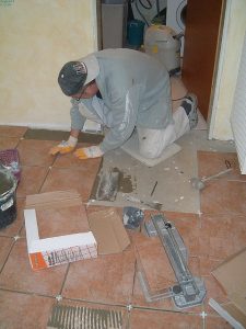 Ein Fliesenleger kniet auf einem teilweise gefliesten Boden und setzt neue Fliesen in den frischen Fliesenkleber ein. Um ihn herum liegen Werkzeug, ein Fliesenschneider, Kartonstücke und Fliesenkreuze zum Ausrichten. Die Szene spielt sich in einem Wohnungsflur ab, im Hintergrund ist eine Waschmaschine sichtbar.