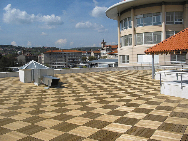 Große Dachterrasse mit im Schachbrettmuster verlegten quadratischen Feinsteinzeugfliesen in Holzoptik, vor einem modernen runden Gebäude mit großen Fenstern.
