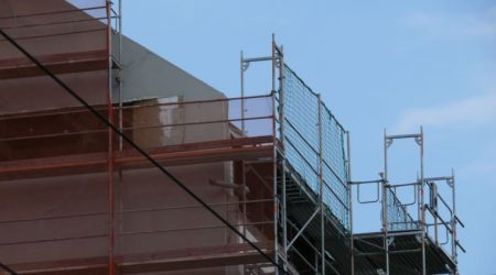 Detailansicht eines Fassadengerüsts mit Schutznetzen und Durchstiegsrahmen an einem mehrstöckigen Gebäude unter blauem Himmel während Bauarbeiten.