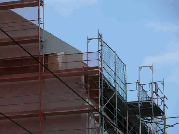 Detailansicht eines Fassadengerüsts mit Schutznetzen und Durchstiegsrahmen an einem mehrstöckigen Gebäude unter blauem Himmel während Bauarbeiten.