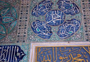 Detailaufnahme kunstvoll verzierter Fliesen in Blau- und Grüntönen mit arabischer Kalligrafie und ornamentalen Mustern an einer Wand, inspiriert von islamischer Architektur.