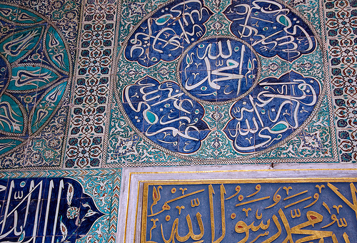 Detailaufnahme kunstvoll verzierter Fliesen in Blau- und Grüntönen mit arabischer Kalligrafie und ornamentalen Mustern an einer Wand, inspiriert von islamischer Architektur.