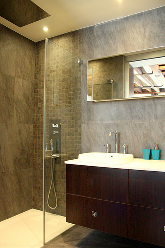 Modernes barrierefreies Badezimmer mit Glasduschabtrennung, dunklen Wandfliesen und wandmontiertem Waschbecken mit großem Spiegel.