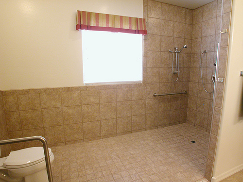 Barrierefreies Badezimmer mit beiger Fliesenverkleidung, bodengleicher Dusche, Haltegriffen und einer Toilette mit zusätzlichem Haltebügel.