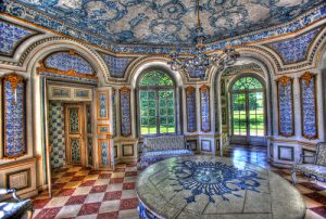 Prunkvoller Barockraum mit blauen Wandfliesen, kunstvoll verziertem Stuck an Wänden und Decke sowie einem großen, runden Tisch mit floraler Intarsie auf dem Marmorfußboden.