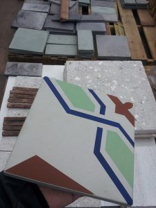 Eine Hand hält eine farbenfrohe Zementfliese mit geometrischem Muster in Grün, Blau, Weiß und Rotbraun, im Hintergrund liegen zahlreiche weitere Fliesen und Platten aus Naturstein oder Zement zur Auswahl auf Paletten.
