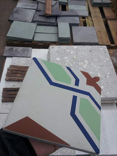 Eine Hand hält eine farbenfrohe Zementfliese mit geometrischem Muster in Grün, Blau, Weiß und Rotbraun, im Hintergrund liegen zahlreiche weitere Fliesen und Platten aus Naturstein oder Zement zur Auswahl auf Paletten.