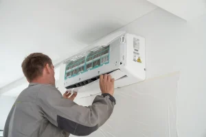 Handwerker in Arbeitskleidung bei der Wartung oder Reinigung einer geöffneten Wand-Klimaanlage in einem weißen Raum.