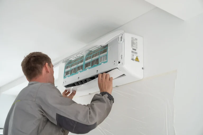 Handwerker in Arbeitskleidung bei der Wartung oder Reinigung einer geöffneten Wand-Klimaanlage in einem weißen Raum.