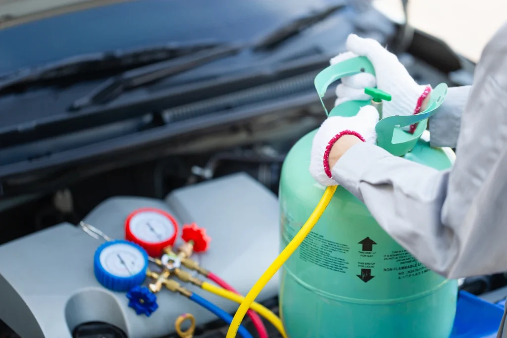 Mechaniker in Handschuhen füllt Kältemittel aus einer hellgrünen Flasche über Manometer-Armaturen in einen Automotor.