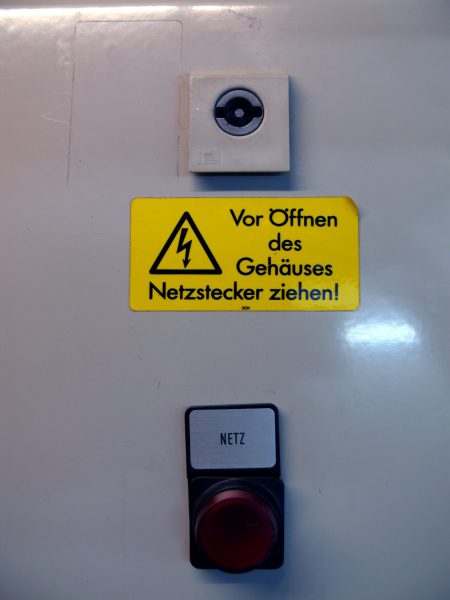 Gehäuse eines elektrischen Geräts mit Warnschild ‚Vor Öffnen des Gehäuses Netzstecker ziehen!‘, darüber eine Netzanschlussbuchse und darunter ein roter Netzschalter.
