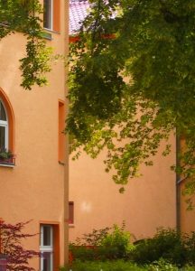 Ein Gebäude mit orangefarbenen Wänden und mehreren Fenstern, umgeben von üppigem Grün. Baumzweige ragen ins Bild und spenden Schatten. Im Vordergrund ist dichte Vegetation zu sehen, wodurch eine ruhige, natürliche Atmosphäre entsteht.