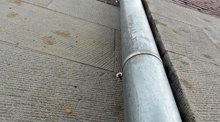 Nahaufnahme einer senkrechten Fallrohrleitung an einer Hauswand. Die Metallrohre verlaufen entlang der Fassade aus Naturstein und Backstein, die durch ihre Struktur und Materialwechsel betont ist. Die Perspektive ist stark nach oben gerichtet.